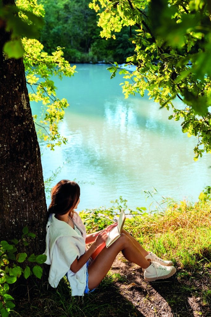 Young woman writing and drawing in notebook sitting under the tree near river on nature in sunlight. Closeness to nature  inspires creativity. Urban escapism.