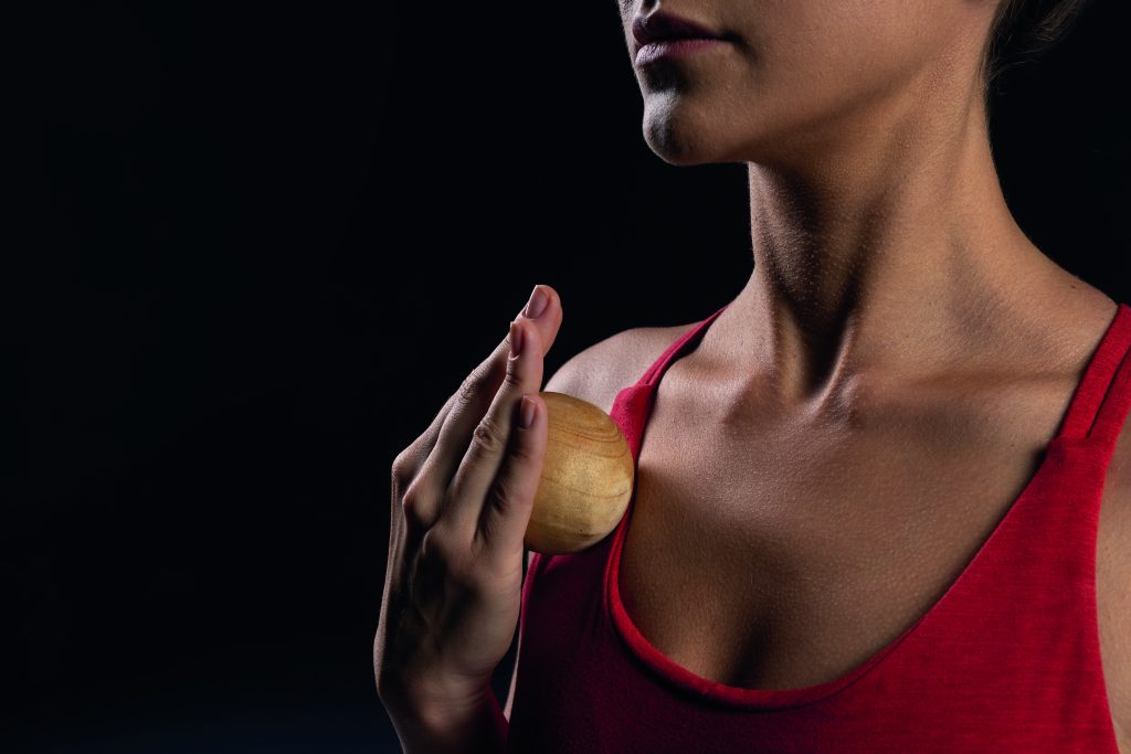 Close up of woman using wooden massage ball on chest wearing red tank top on dark background, self care and relaxation techniques. self massage tool