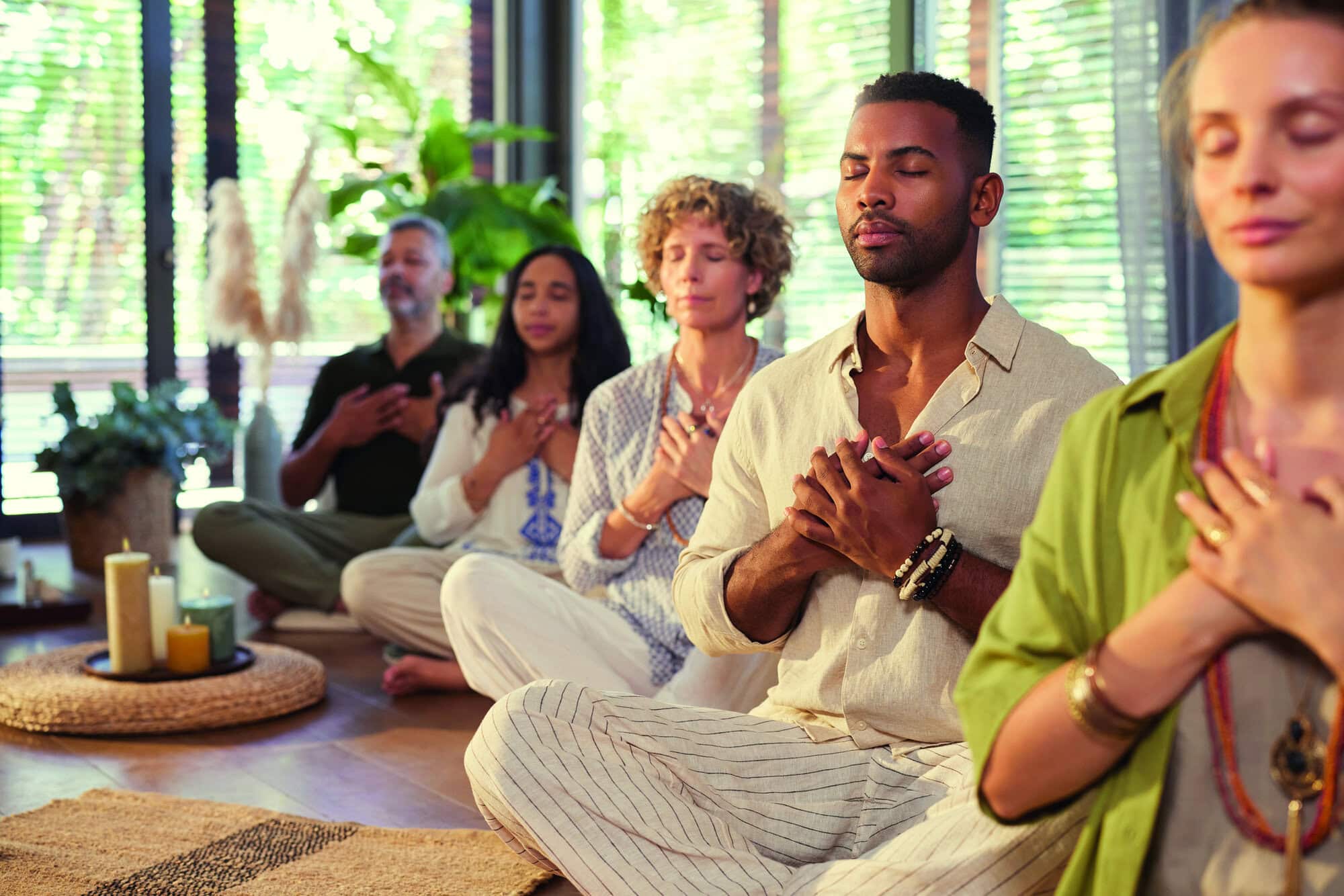 Diverse group meditating with hands on heart at indoor wellness retreat with copy space. Group of people sitting together in a line practicing mindfulness meditation with hand on chest and closed eyes. Peaceful session focused on gratitude, healing, and connection.