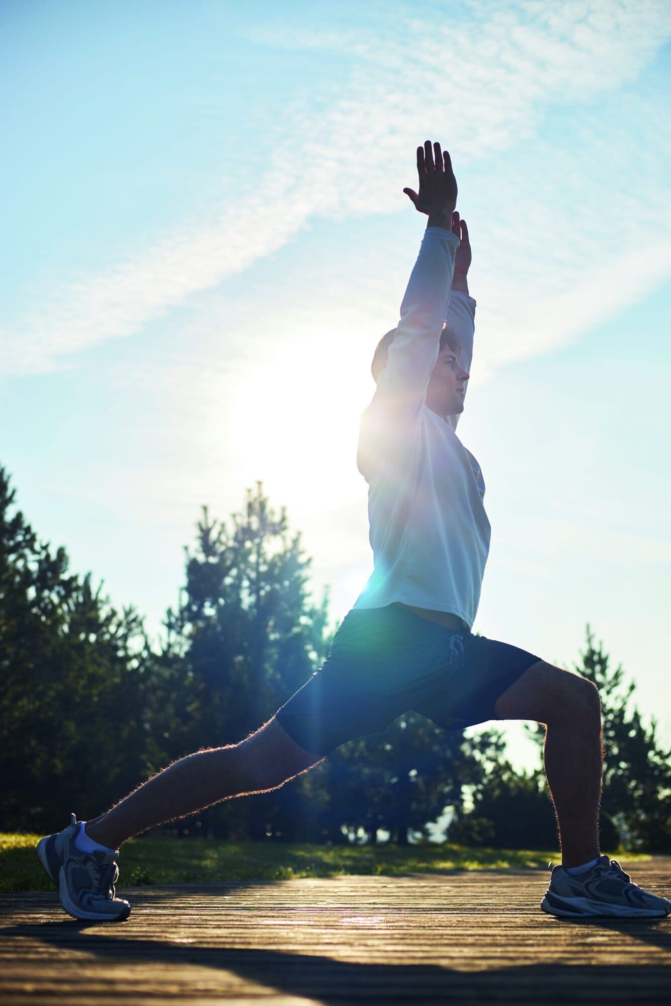 Young adult Caucasian man performing outdoor yoga stretching exercise on wooden platform with arms raised overhead, sunlight shining through trees in background, wearing athletic shoes