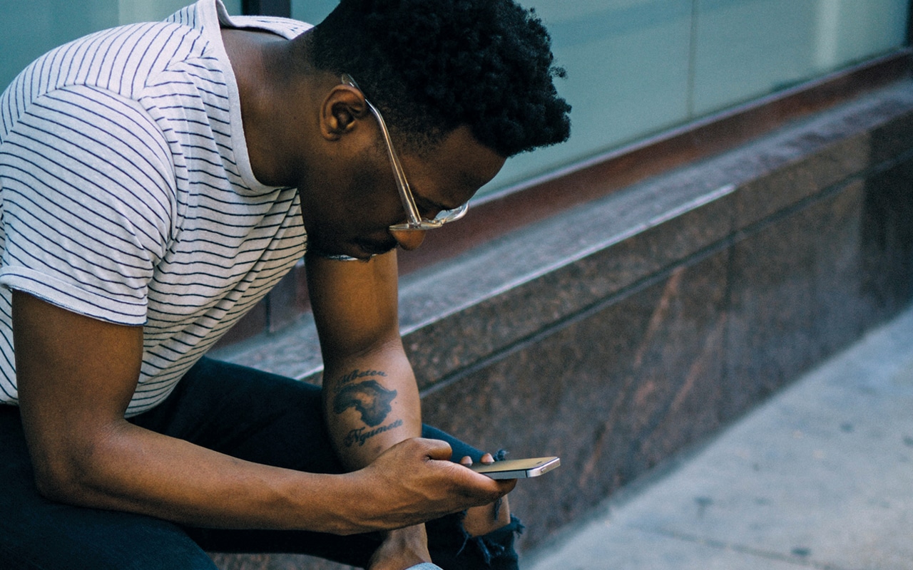 A man sits outside on a wall, he has sunglasses on and a tattoo on his lower left arm. He is holding a phone in his right hand, and looking down at it. 