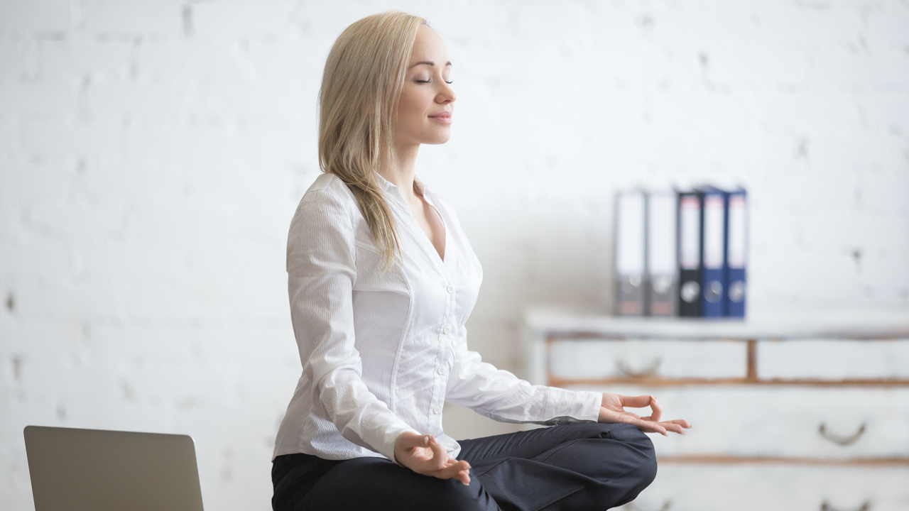 A woman in office wear - a white shirt and dark trouser - is sitting in a meditation pose on a desk with office stuff around her. 