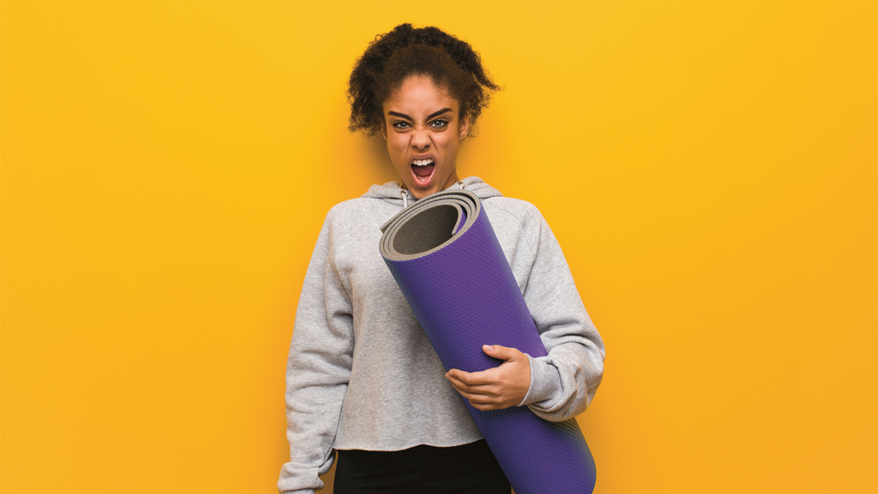 A woman standing against a yellow background: she holds a purple yoga mat and is screaming at the camera. 