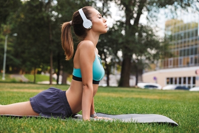 Fit young sportswoman dressed in sportswear listening to music with headphones while doing stretching exercises on fitness mat on grass outdoors