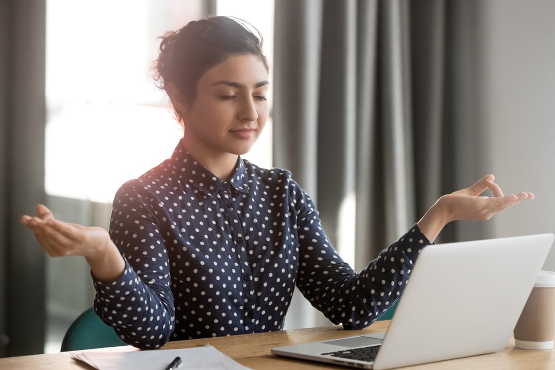 Mindful calm indian young business woman meditate at work desk with eyes closed, healthy hindu girl take break relax in office doing yoga at workplace feel balance no stress and peace of mind concept