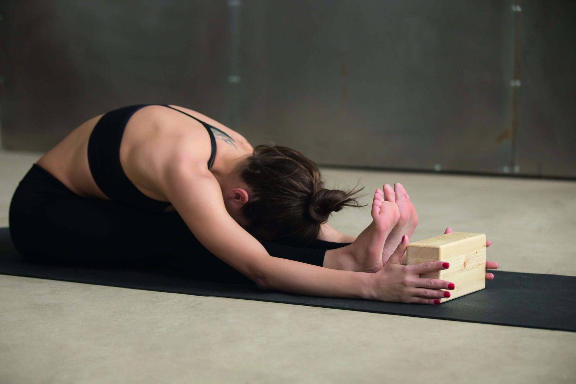 Young attractive woman practicing yoga, stretching in paschimottanasana exercise, using wooden block, Seated forward bend pose, wearing sportswear, working out, full length, cool urban style, studio