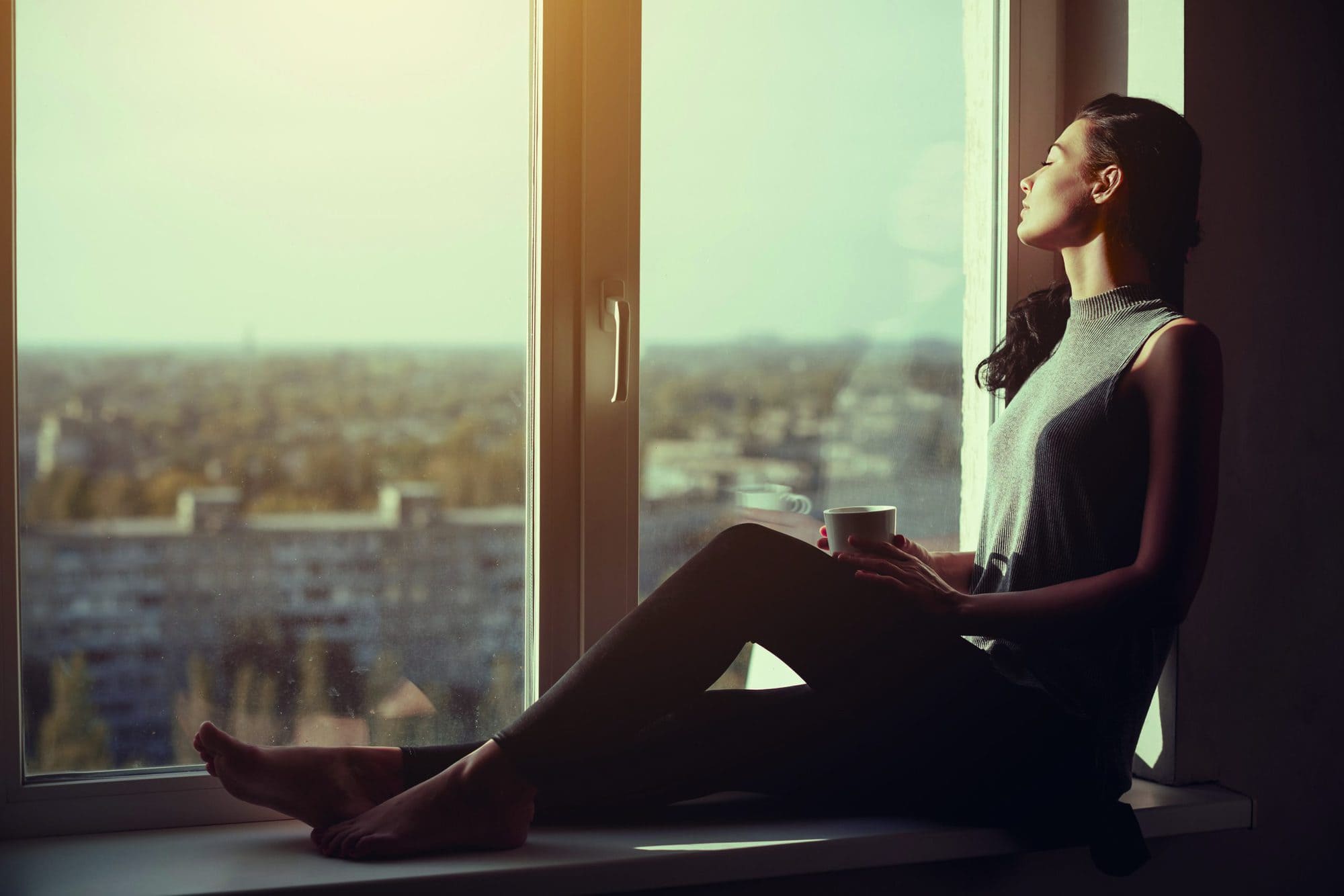 Resting and thinking woman with closed eyes. Calm girl with cup of tea or coffee sitting on the window-sill at home. Side view. Young attractive multi-racial Asian Caucasian lady in casual clothes