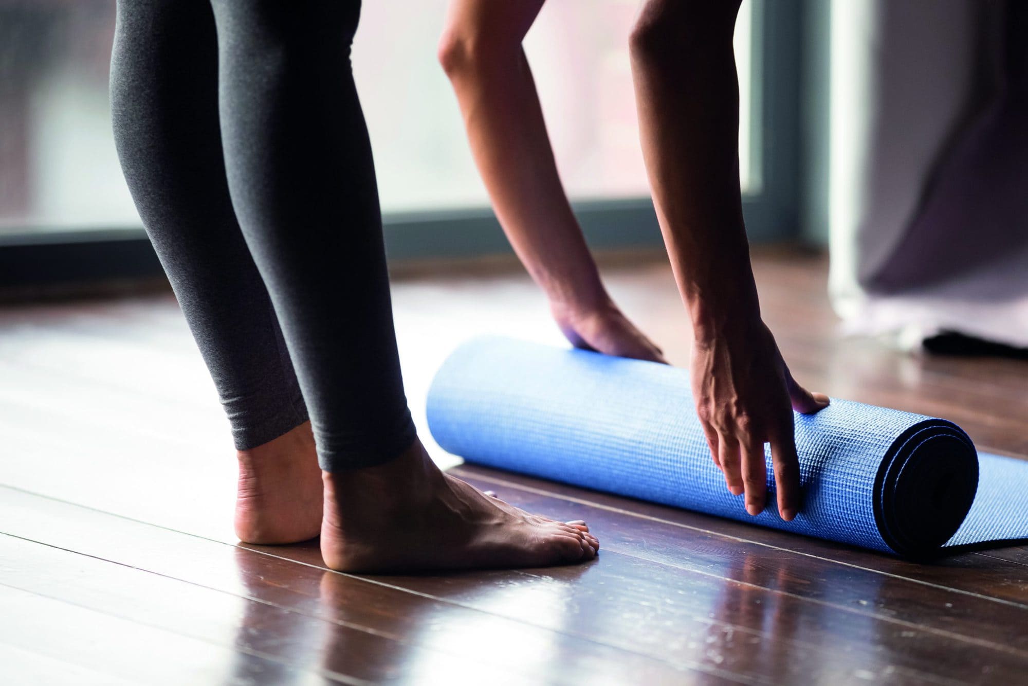 Time for meditation, fitness session, well-being concept. Girl wearing grey sporty pants rolling fitness mat before, after class in yoga studio club or at home on wooden floor. Hands and legs close up