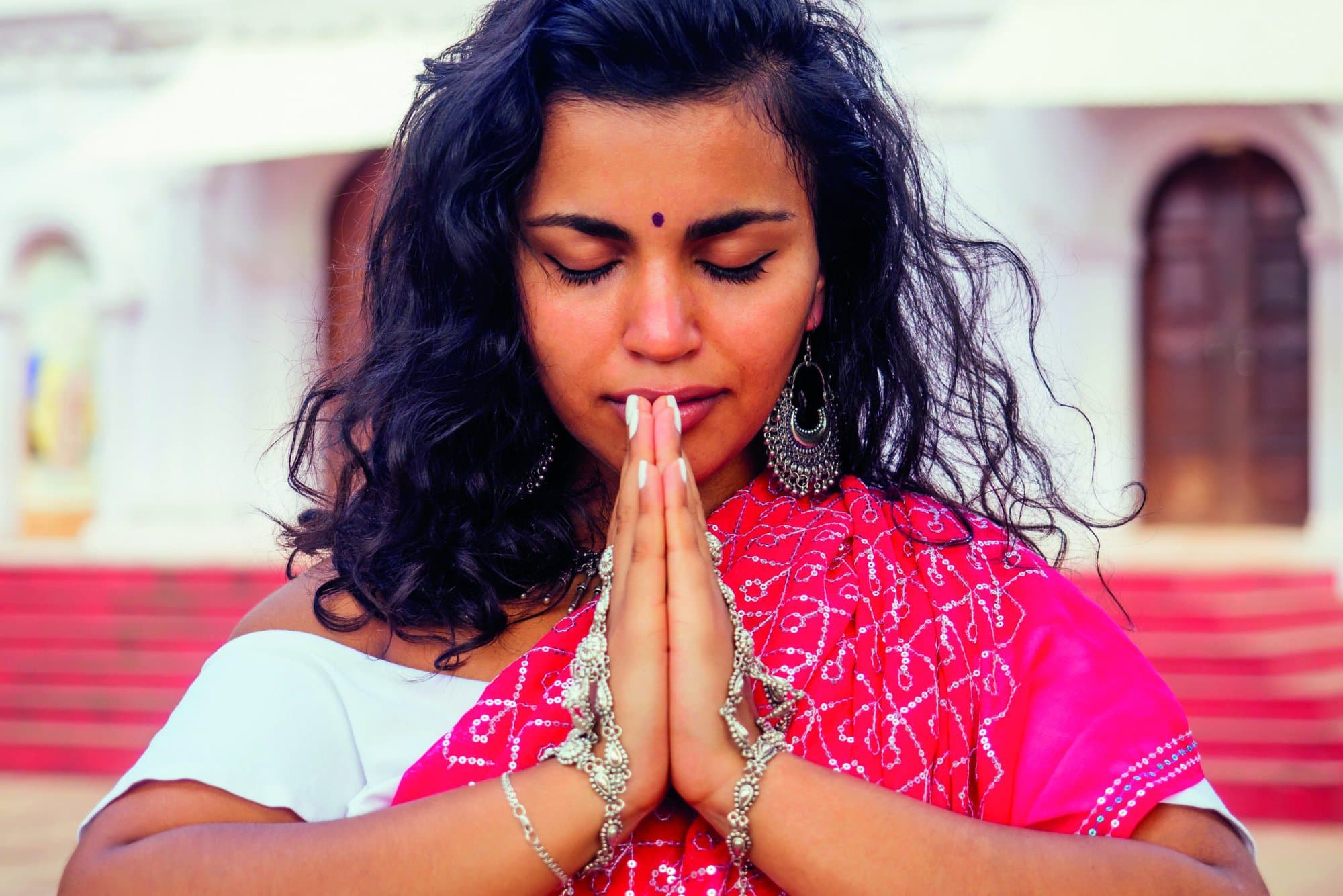 Young Indian woman in traditional sari red dress praying in a hindu temple goa india Hinduism.girl performing namaste gesture catholicism Delhi Street holi festival.om yoga meditation female model