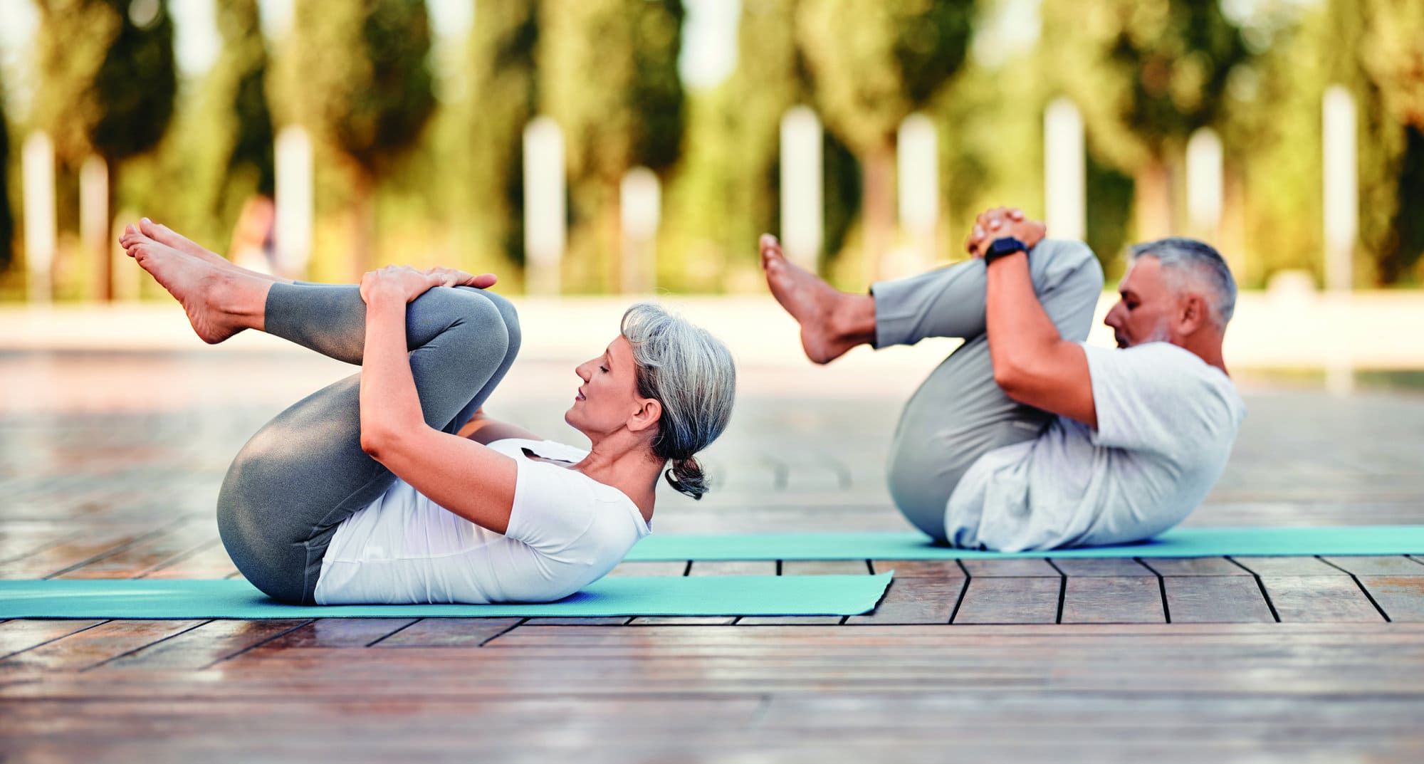 Active mature family man and woman doing yoga exercise  lying on mat hugging her knees to chest after yoga practice outdoor in park, healthy active senior couple doing stretching exercises