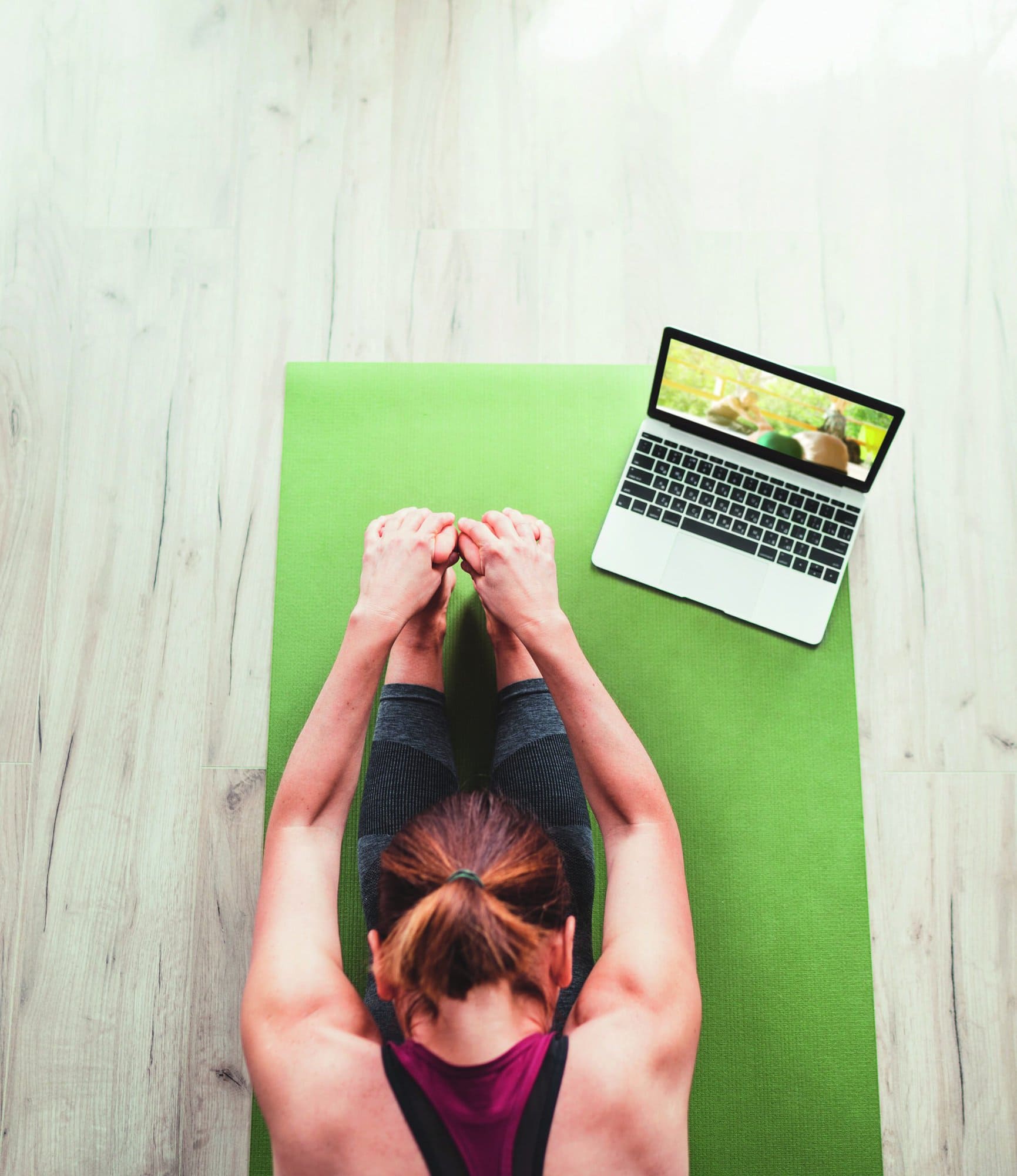 Top view at fit sporty healthy woman sitting on mat in Paschimottanasana pose, doing breathing exercises, watching online yoga class on laptop computer. Healthy people and self motivation concept.