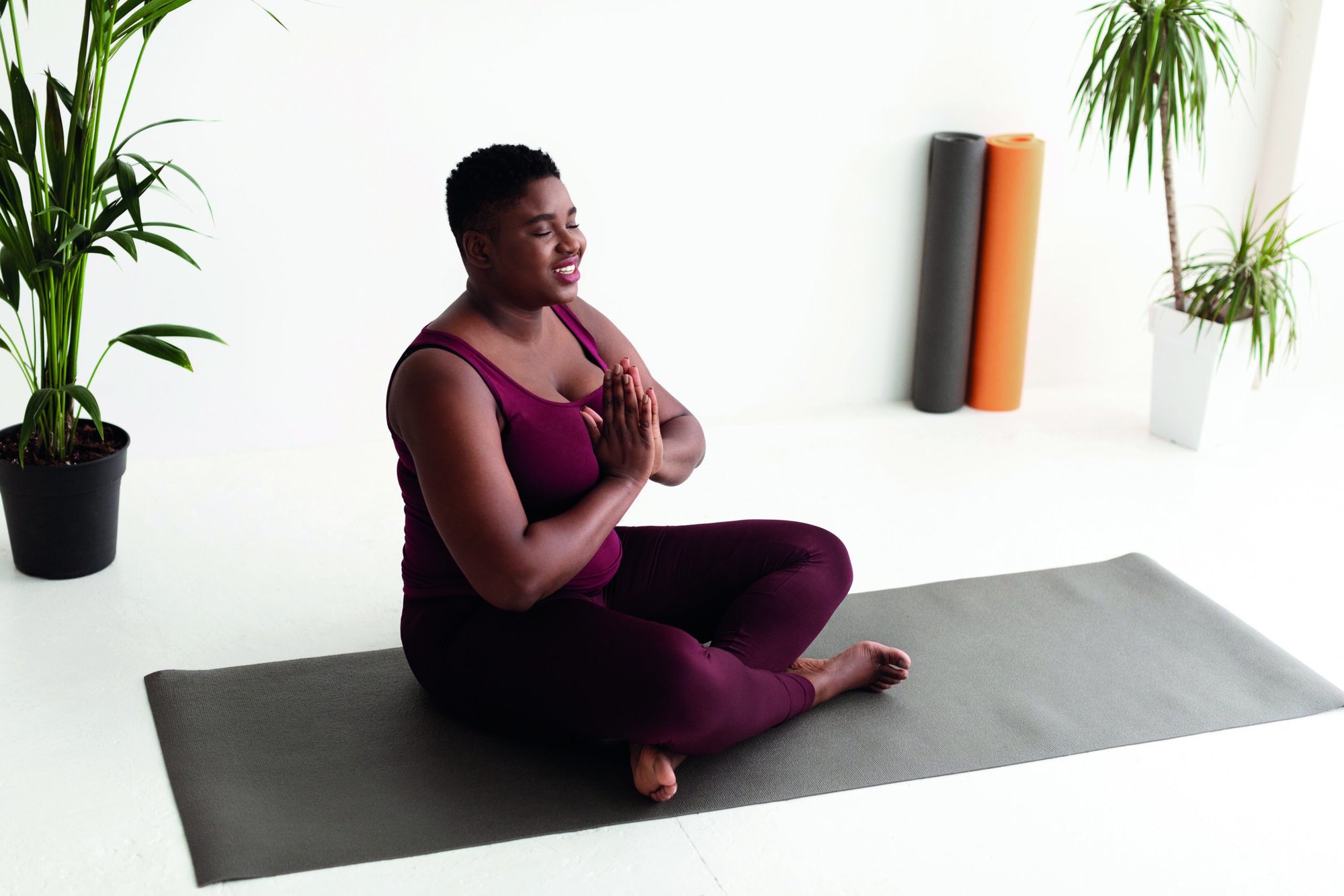 Relaxed overweight short-haired young african american woman in sportswear meditating with closed eyes at yoga studio, holding hands in namaste gesture and smiling, copy space