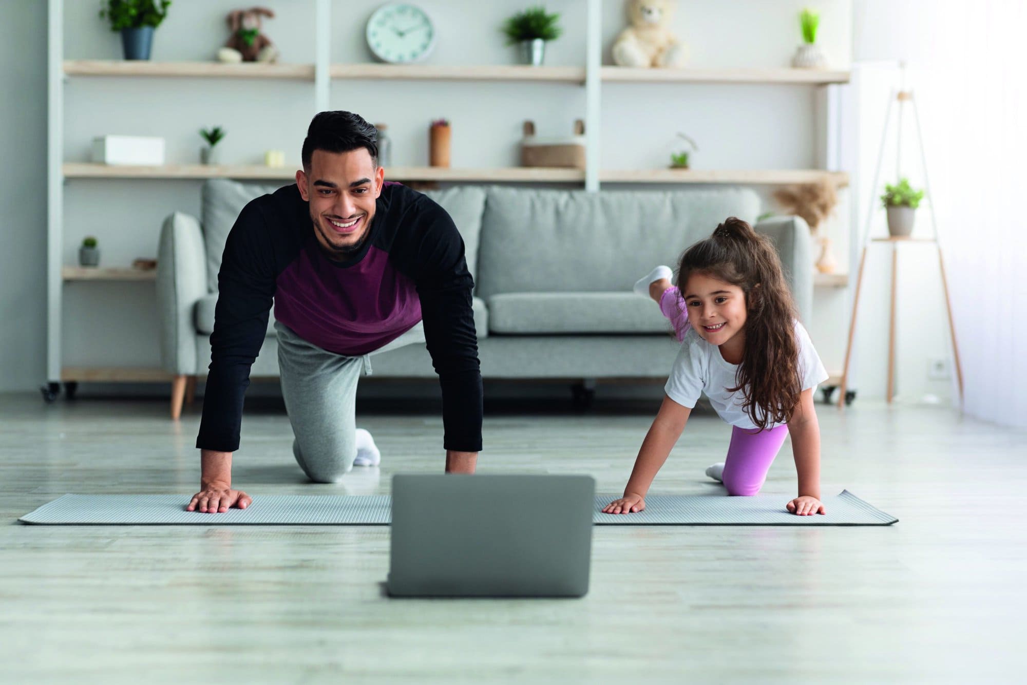 Happy arab father and little daughter having online fitness class, exercising at home together, wearing sportswear, training on yoga mat in front of laptop, enjoying workout, copy space