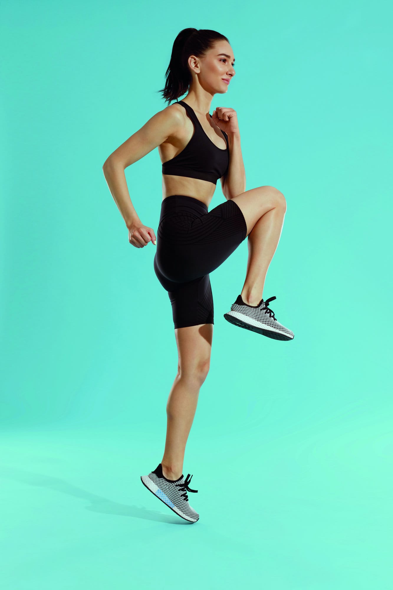 Fitness workout. Sport woman exercising, jumping on blue background. Full length portrait of sporty girl in black sportswear running on spot, warming up at studio