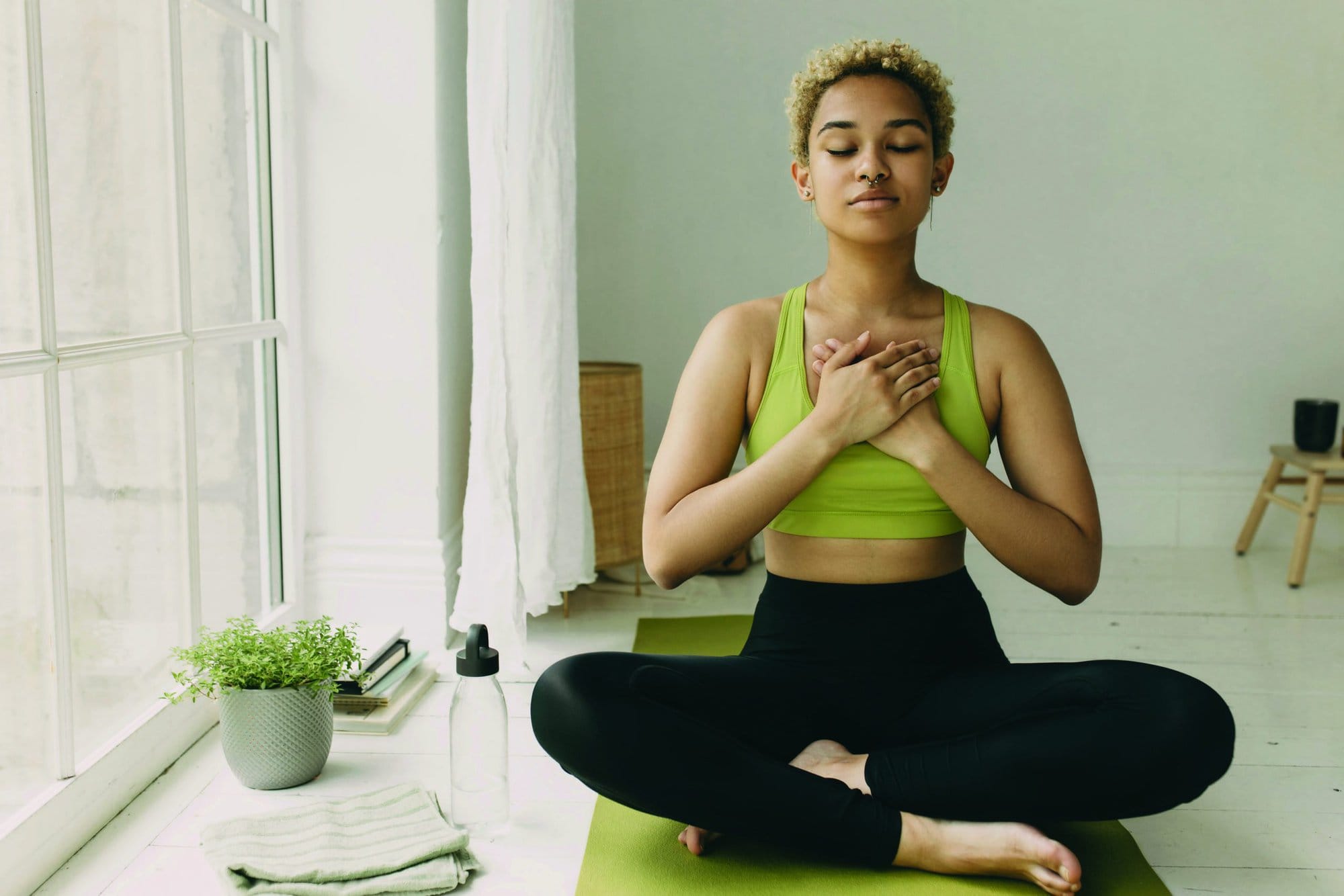 Young African American lady practicing yoga at home in black leggings sitting on floor in lotus posture hands crossed on chest slight delight smile on face, feeling her body. Emotional health