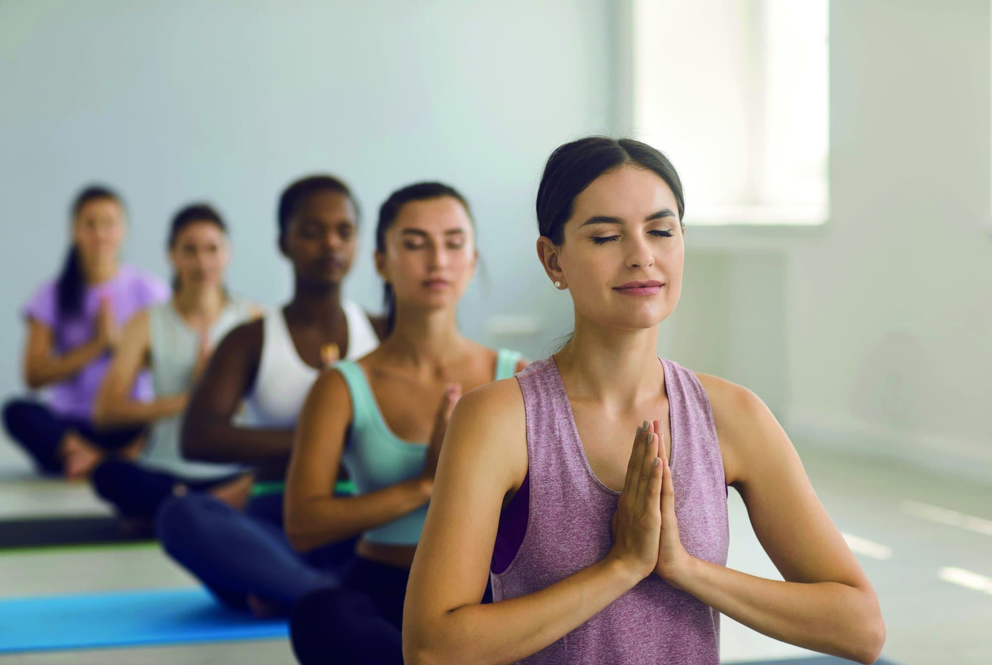 Portrait of a relaxed athletic woman enjoying meditation with closed eyes at a yoga class at a sports club. Woman sits in a lotus position with folded palms in a group yoga class. Blurred background.