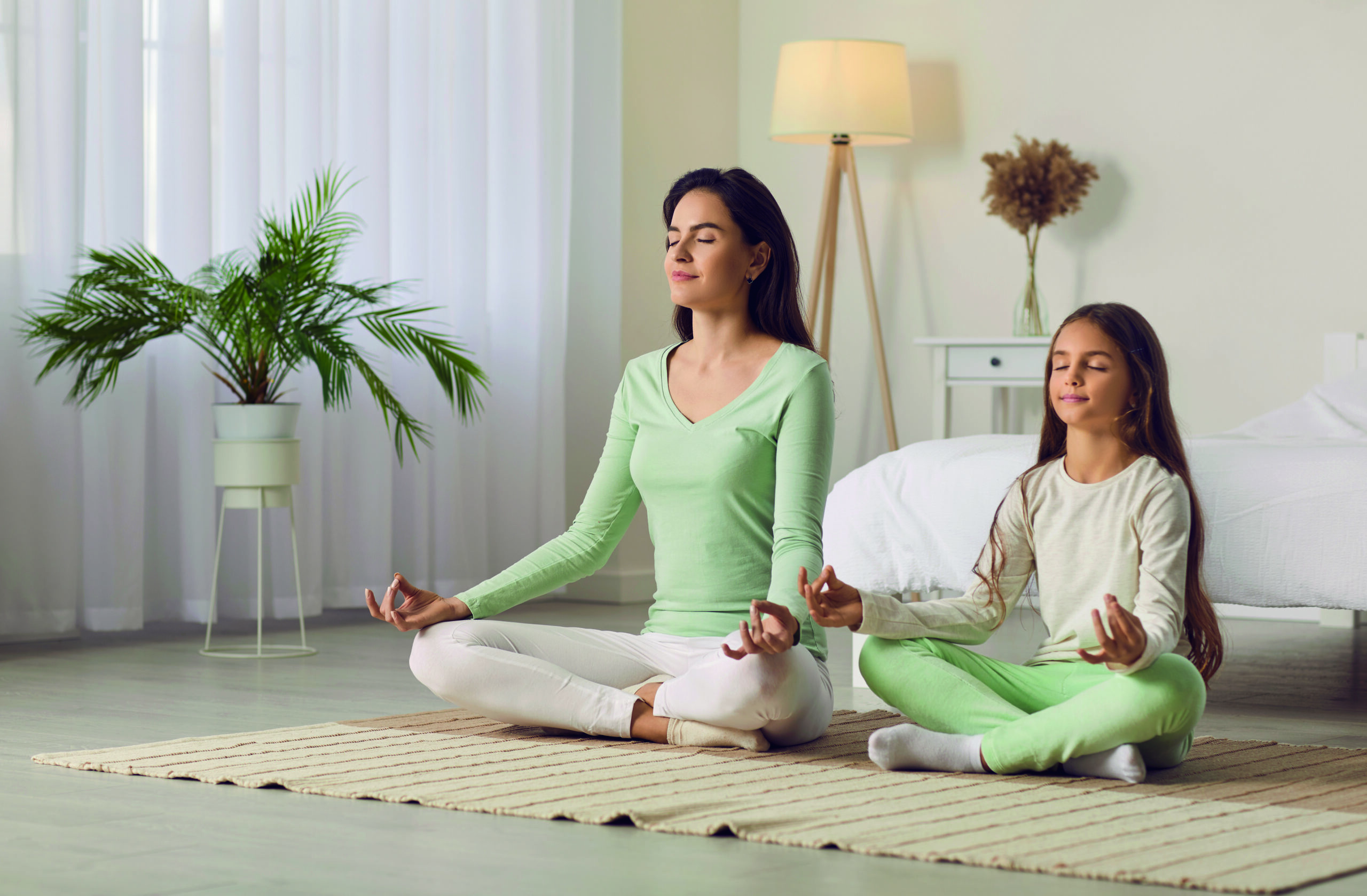 Family yoga. Beautiful young woman and her charming little daughter are smiling while doing yoga together at home. Family sits in lotus position on floor in living room. Mom teaches child to meditate.