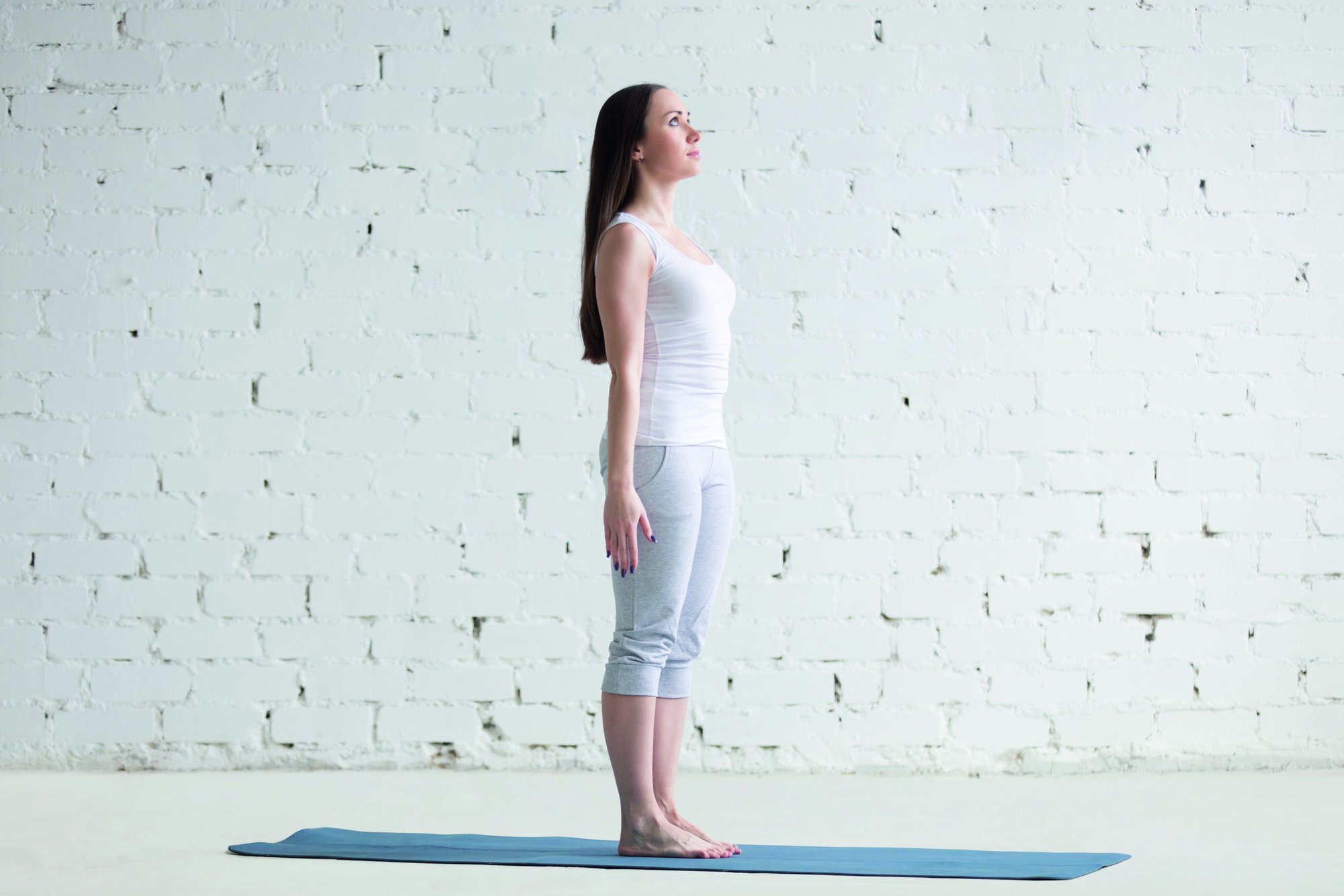 Attractive happy young woman working out indoors. Side view portrait of beautiful model doing yoga exercise on blue mat. Standing in Tadasana, Mountain Pose. Full length