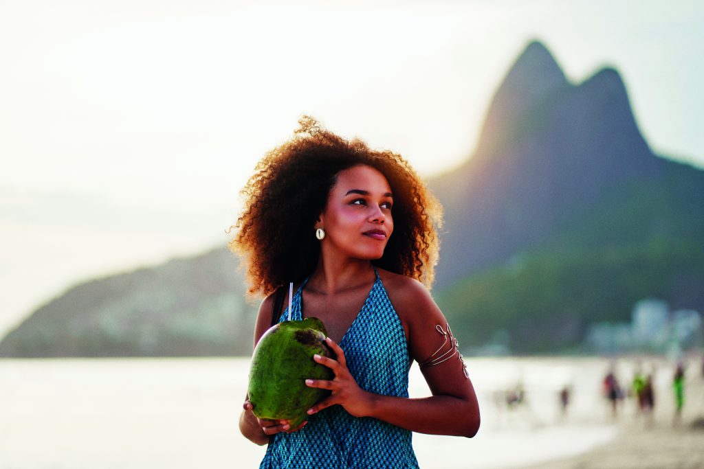 waist up portrait cheerful young brazilian afro hairstyle woman walking on the beach holding a coconut water in Ipanema
