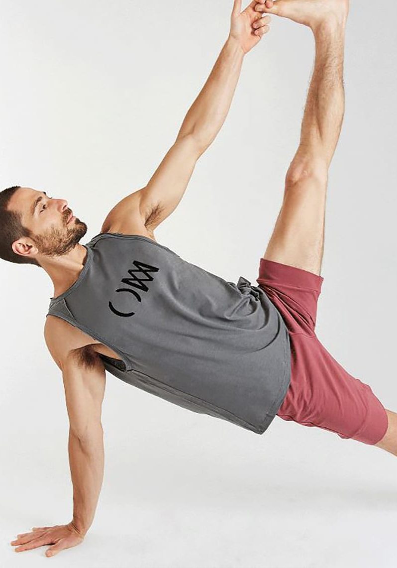 A man is balancing on his right arm and right leg; his left arm is extended into the air and his left hand is clasping his left foot, which is also extended into the air. He is wearing a grey t-shirt and light red shorts. 