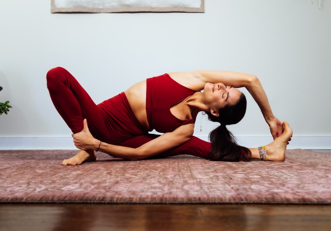 Elena Brower in a yoga pose - she is seated, stretching her body to the left, with her right arm over her head and touching her left foot, with her right leg bent at the knee, her foot on the floor, and her left hand holding her lower right leg. 