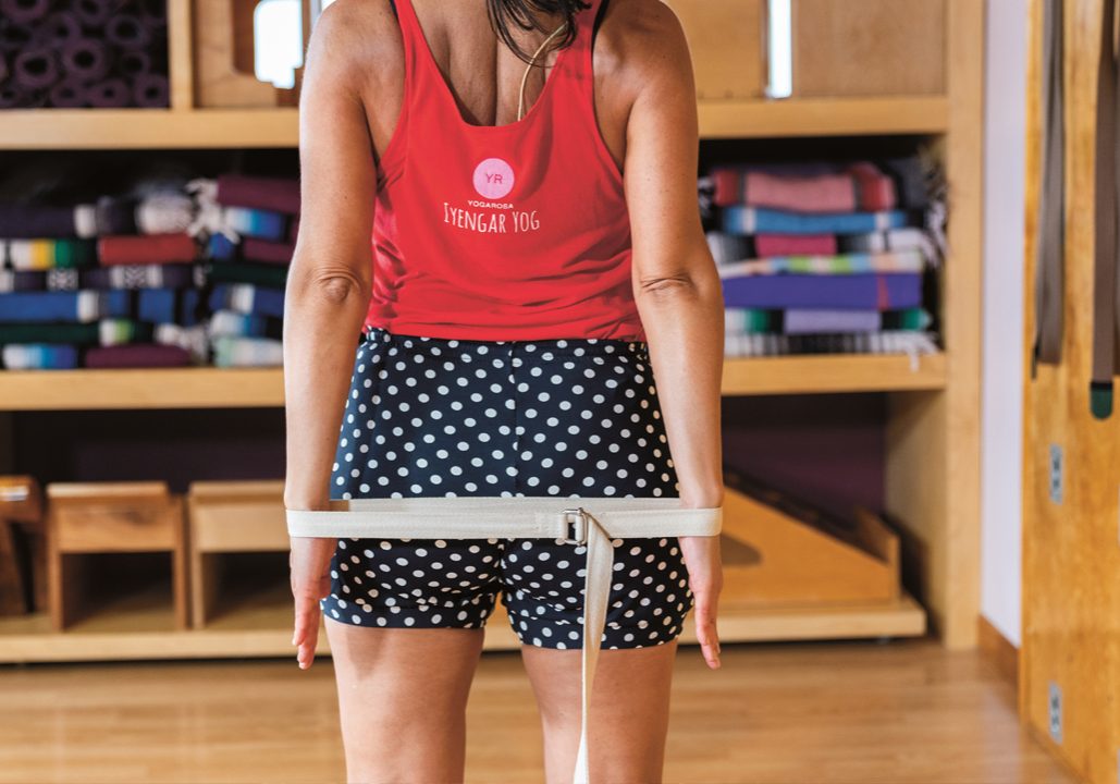 A woman faces away from the camera, her hands are at her side slightly behind her back. A yoga belt is looped around her wrists. 