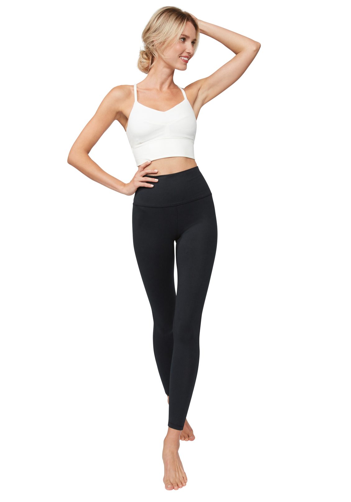 A woman stands with her right hand on her hip, and her left arm bent at the elbow with her hand on her head. Her left foot is slightly in front of her right foot. She is wearing a white cropped top and a pair of black high waisted leggings.
