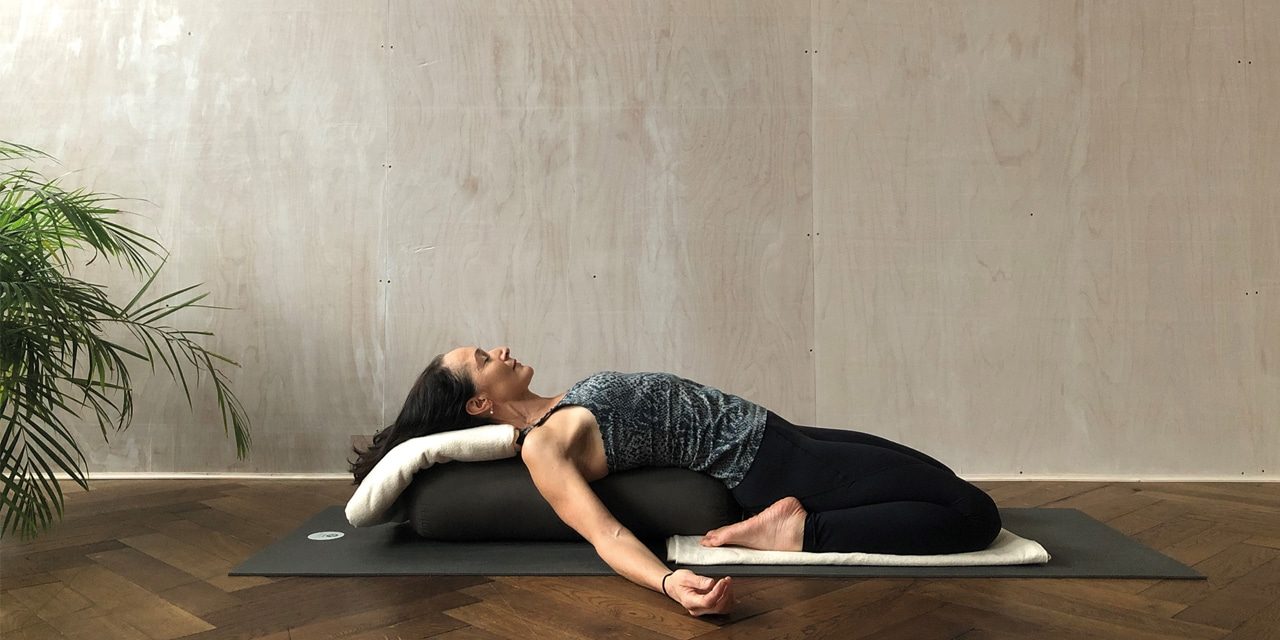 A woman lays on a yoga mat and bolsters with her legs bent beneath her and her arms splayed by her side. She has her eyes closed. 