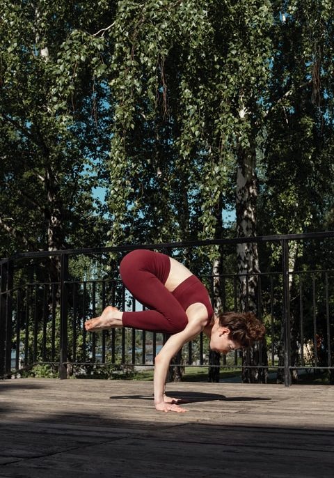 Young brunette woman practicing Bakasana crow pose in the park on wooden platform, selective focus