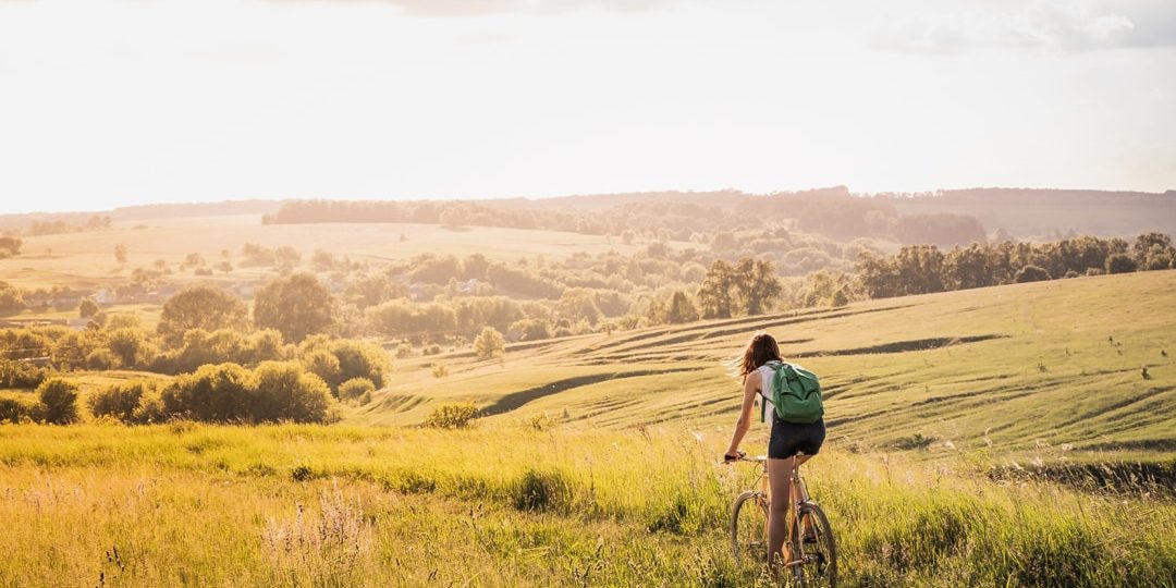 Girl riding a bicycle down the hill in beautiful rural landscape at sunset. Young pretty female person with retro bike standing in a meadow on bright sunny afternoon in summer