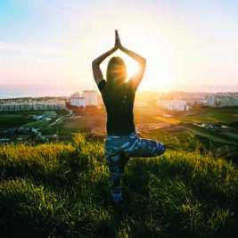 Woman doing yoga on the green grass on the top of the mountain with beautiful view at sunset or sunrise. Tree pose or vrksasana with hands together up above head. Relaxation, harmony with nature