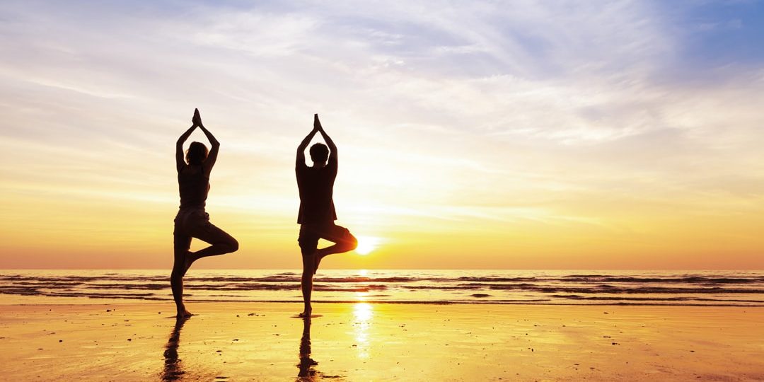 Two,People,Practicing,Yoga,Tree,Position,On,The,Beach,With Two people practicing yoga tree position on the beach with beautiful sunset and reflection