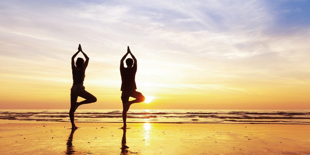 Two people practicing yoga tree position on the beach with beautiful sunset and reflection